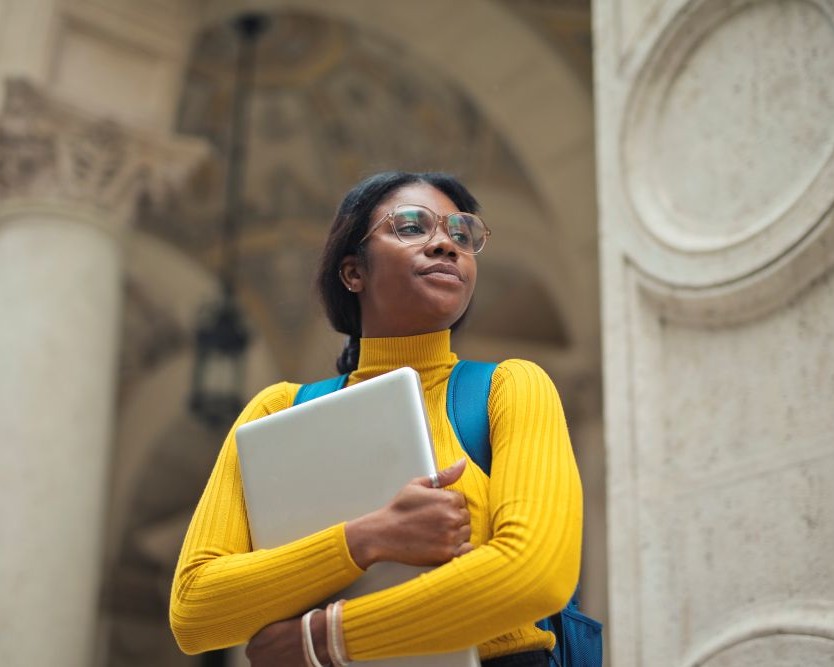 Student in a yellow sweater holding a laptop outside a university building